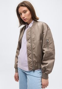 Tânără purtând o jachetă bomber matlasată taupe, un tricou polo lavandă și blugi deschiși, stând în picioare pe un fundal simplu.