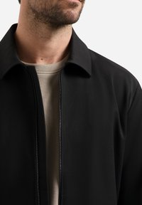 Mann in schwarzer Zip-Up-Jacke über einem beigefarbenen Rundhals-Shirt, der Untergesicht und Hals vor einem weißen Hintergrund zeigt.