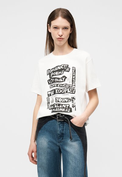 Γυναίκα που φοράει λευκό γραφικό t-shirt με μαύρο κείμενο και ψηλόμεσα τζιν σε δύο αποχρώσεις, μπλε και μαύρο, στέκεται με το ένα χέρι στην τσέπη.