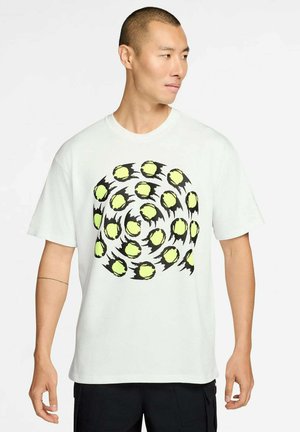 Mann trägt weißes T-Shirt mit einem spiralförmigen Muster aus gelben und schwarzen Tennisbällen, schaut nach rechts vor einem einfarbigen Hintergrund.