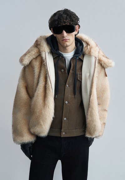 Manteau surdimensionné en fausse fourrure couleur beige clair, avec capuche, fermeture éclair à l'avant et doublure crème. Porté avec une veste en denim marron foncé et un pantalon foncé.