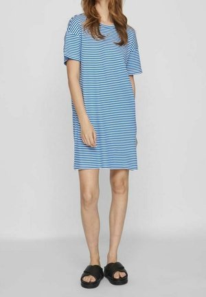 Vrouw draagt een blauw-wit gestreepte korte mouw jurk met zwarte slippers, staand tegen een effen lichte achtergrond.