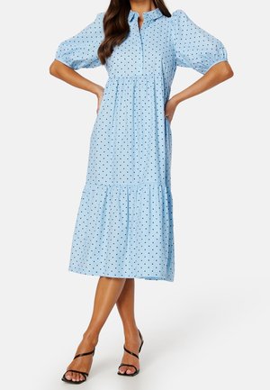 Femme portant une robe midi bleu clair à pois noirs, manches bouffantes, col boutonné et talons noirs à lanières.