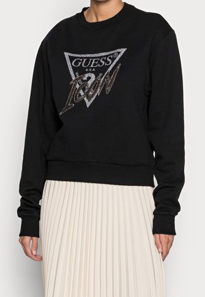 Czarna bluza z długim rękawem z dużym srebrnym logo "Guess Icon" z przodu, zestawiona z białą plisowaną spódnicą z wysokim stanem.