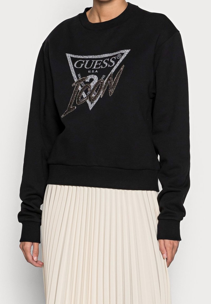 Sweat-shirt noir à manches longues avec un grand logo argenté « Guess Icon » sur le devant, associé à une jupe plissée blanche taille haute.