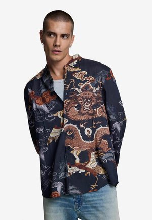 Uomo che indossa una camicia scura con dettagliati disegni di drago e gru, abbinata a jeans azzurri chiari, guarda di lato su uno sfondo bianco.