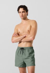 Zelené plavkové šortky z ľahkej tkaniny s elastickým pásom a čiernym sťahovacím šnúrkou. Na boku majú jednu detailnú logo aplikáciu.