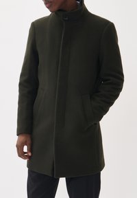 Manteau en laine vert olive avec un col montant, deux poches avant et un design sans couture. Texture douce, coupe ajustée et détail de fermeture à boutons.