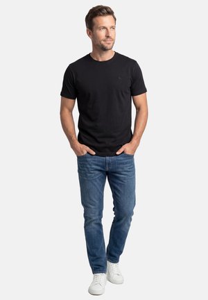 Mand iført sort T-shirt med korte ærmer, blå jeans, hvide sneakers, hænderne i lommerne, stående mod en ensartet lys baggrund.