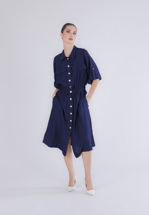 Nainen tummansinisen, napitettavan midi-mekon kanssa, jossa on käärityt hihat ja joustava vyötärö, seisoo käsivarret taskuissa, valkoiset korkokengät jalassa.