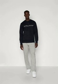 Σκούρο ναυτικό φούτερ με λογότυπο "EJÖRN BORG" σε ανοιχτό γκρι, συνδυασμένο με ανοιχτό γκρι παντελόνια και άσπρα αθλητικά παπούτσια, σε στάση μπροστά από μια απλή γκάμα.