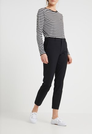 Femme portant un pantalon noir ajusté, un t-shirt à manches longues rayé noir et blanc, et des baskets blanches à lacets, debout devant un fond uni.