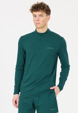 Jonge man met krullend haar, gekleed in een donkergroen sportshirt met lange mouwen en bijpassende short, staat met één hand in zijn zak.
