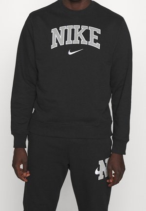 Mand iført sort Nike sweatshirt med stort logo på brystet og matchende sorte bukser med mindre logo på låret.