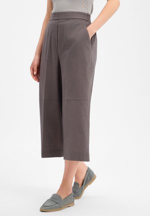 Frau trägt weite graue kurze Hosen mit elastischem Bund und graue Slip-on-Loafer und steht vor einem einfarbigen Hintergrund.