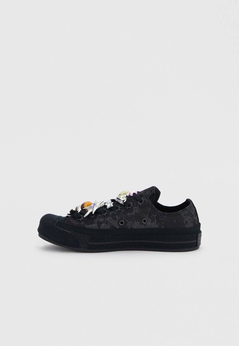 Black low-top sneaker with subtle floral pattern and decorative silver and amber charms attached to the side and laces on a white background.