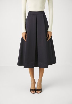 Femme portant une jupe midi noire avec un pli sur le devant, associée à un haut blanc à manches longues et des talons ouverts marron, debout devant un fond blanc.