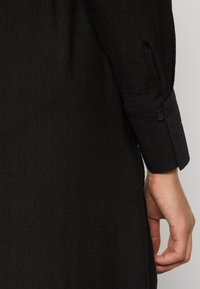 Μαύρο πουκάμισο από ύφασμα με υφή. Το μανίκι διαθέτει κουμπί κλεισίματος και μια κοπτή λεπτομέρεια κοντά στην περιοχή του καρπού.