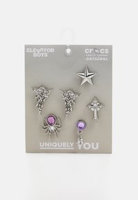 Conjunto de cinco charms metálicos: dos cabezas de dragón, una estrella, una cruz y una araña con acentos morados, montados en una tarjeta gris etiquetada como "Únicamente Tú."