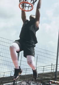 Čierny športový top, čierne šortky a biele legíny s logom Jordan. Dunkuje basketbal do oranžového koša proti zamračenému nebu.