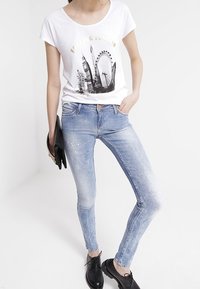 Mujer vestida con una camiseta gráfica blanca con tema de Londres, jeans ajustados de color azul claro con salpicaduras de pintura, zapatos negros y sosteniendo un clutch negro.