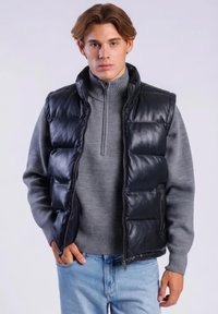 Jonge man draagt een zwarte gewatteerde bodywarmer over een grijze trui met rits en lichtblauwe spijkerbroek, staand met één hand in zijn zak.