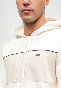 Φούτερ με κουκούλα σε κρέμα και μπεζ, διαθέτοντας μπροστινό φερμουάρ, αντιθετική μαύρη ρίγα και διακριτό πράσινο κεντημένο λογότυπο στο στήθος.