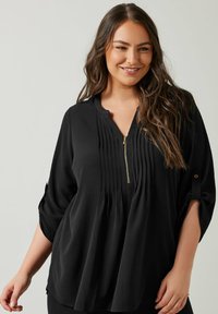 Sort bluse lavet af blødt stof, med plisseret front, lynlås lukning og trekvartærmer med mulighed for at rulle op og guldknap detaljer.