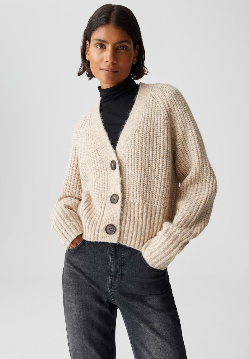 Donna che indossa un cardigan spesso beige con grandi bottoni su un dolcevita nero e jeans grigio scuro, con le mani in tasca, rivolta in avanti.