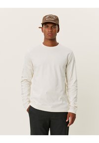 Pitkähihainen kerma-valkoinen T-paita, joka on valmistettu puuvillasta ja jossa on graafisia printtejä molemmissa hihoissa. Yhdistettynä ruskeaan lippikseen, jossa on brodeerattu teksti.