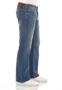 Jeans de mezclilla azul con un corte relajado, lavado deslavado y dos bolsillos frontales. Presenta un parche de cuero marrón en la cinturilla. Zapatillas blancas visibles.