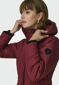 Veste matelassée bordeaux avec capuche, présentant des accents de poignet noirs et un patch logo rond sur la manche. Texture lisse et fermeture à boutons.