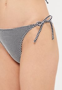 Melnas un baltas šaha uzzīmētas bikini apakšdaļas ar maigu, teksturētu audumu, kuru sānos rotā saišu aizdares. Apmales malas uzsver dizainu.