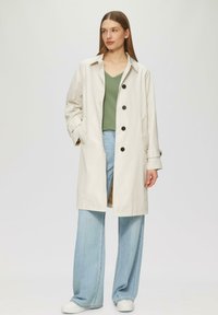 Helles beiges Trenchcoat mit einem Kragen und sechs dunklen Knöpfen, kombiniert mit einem grünen V-Ausschnitt-Oberteil und weit geschnittenen hellblauen Jeans. Weiße Sneakers.