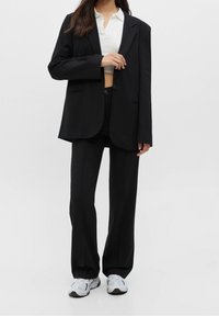Black oversized blazer with two front pockets, paired with fitted black pants and a white cropped polo shirt. White sneakers complete the look.