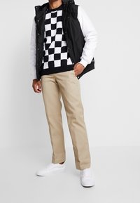 Homme portant un pantalon beige, des baskets blanches, un pull à carreaux noir et blanc, et un gilet sans manches noir avec des manches blanches, se tenant avec une main dans sa poche.