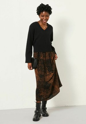 Frau trägt einen schwarzen V-Ausschnitt-Pullover, einen braun-schwarz gemusterten Midirock, schwarze Schnürstiefel, hält eine schwarze Clutch und lächelt, während sie nach unten schaut.