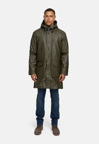 Olijfgroene waterdichte parka met een capuchon, twee voorzakken, verstelbare trekkoorden en een rechte snit. Gedragen over blauwe jeans en bruine schoenen.