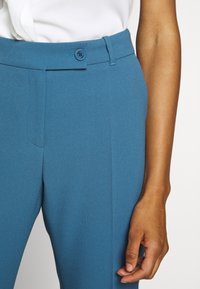 Gros plan d'une personne portant un pantalon bleu texturé avec une taille boutonnée et un haut blanc rentré, main détendue sur le côté.