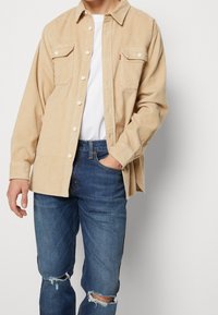 Beige kordflosskjorta med knappstängning, två bröstfickor och en avslappnad passform, ihop med blå slitna jeans.