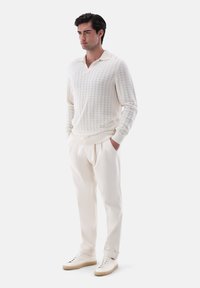 Mann steht mit den Händen in den Taschen, trägt einen cremfarbenen Strickpullover, passende gefaltete Hose und weiße Sneakers mit beigen Sohlen.