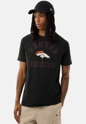 Person mit langem geflochtenem Haar, die eine schwarze Kappe, ein schwarzes Denver Broncos T-Shirt und beige Hose trägt, steht mit einer Hand in der Tasche.