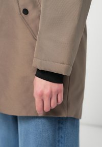 Bež plašč z teksturirano površino, ki ima črn manšeti pod rokavom. Rokav se konča tik nad zapestjem, z rahlim šivom.