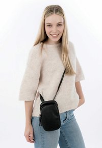 Schwarze Leder-Crossbody-Tasche mit strukturierten Oberfläche, rechteckiger Form und verstellbarem Tragegurt, getragen über einem hellen, fusseligen Pullover.