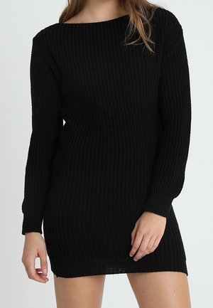 Woman wearing a black, long-sleeve, ribbed knit dress standing against a plain light background.