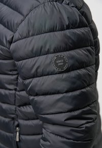 Temno modra napihnjena jakna s horizontalno prešito teksturo. Ima okroglo črno logotipno nalepko na levem rokavu. Vidna podrobnost zadrge.