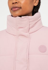 Světle růžová prošívaná bunda s vysokým límcem, prošívanou texturou a kruhovou nášivkou s logem značky na levé straně.