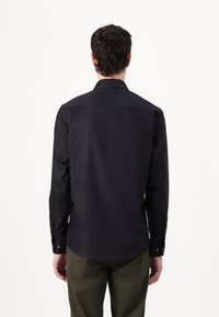 Long-sleeve dark navy shirt made of smooth fabric, featuring a classic collar and buttoned cuffs, worn with olive green pants.