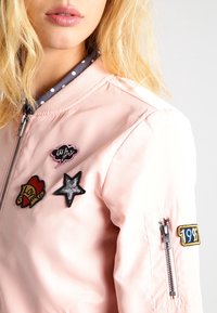 Różowa kurtka typu bomber z naszywkami: "LOVE", "WHY" oraz gwiazdką; zamek błyskawiczny na lewej rękawie; kołnierz w groszki; gładka faktura.
