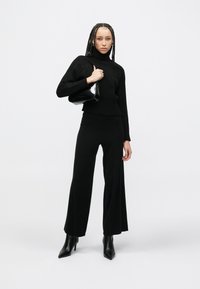 Crni pleteni džemper s visokim ovratnikom, upareni s crnim širokim hlačama. Model drži malu crnu torbicu i nosi šiljaste čizme.